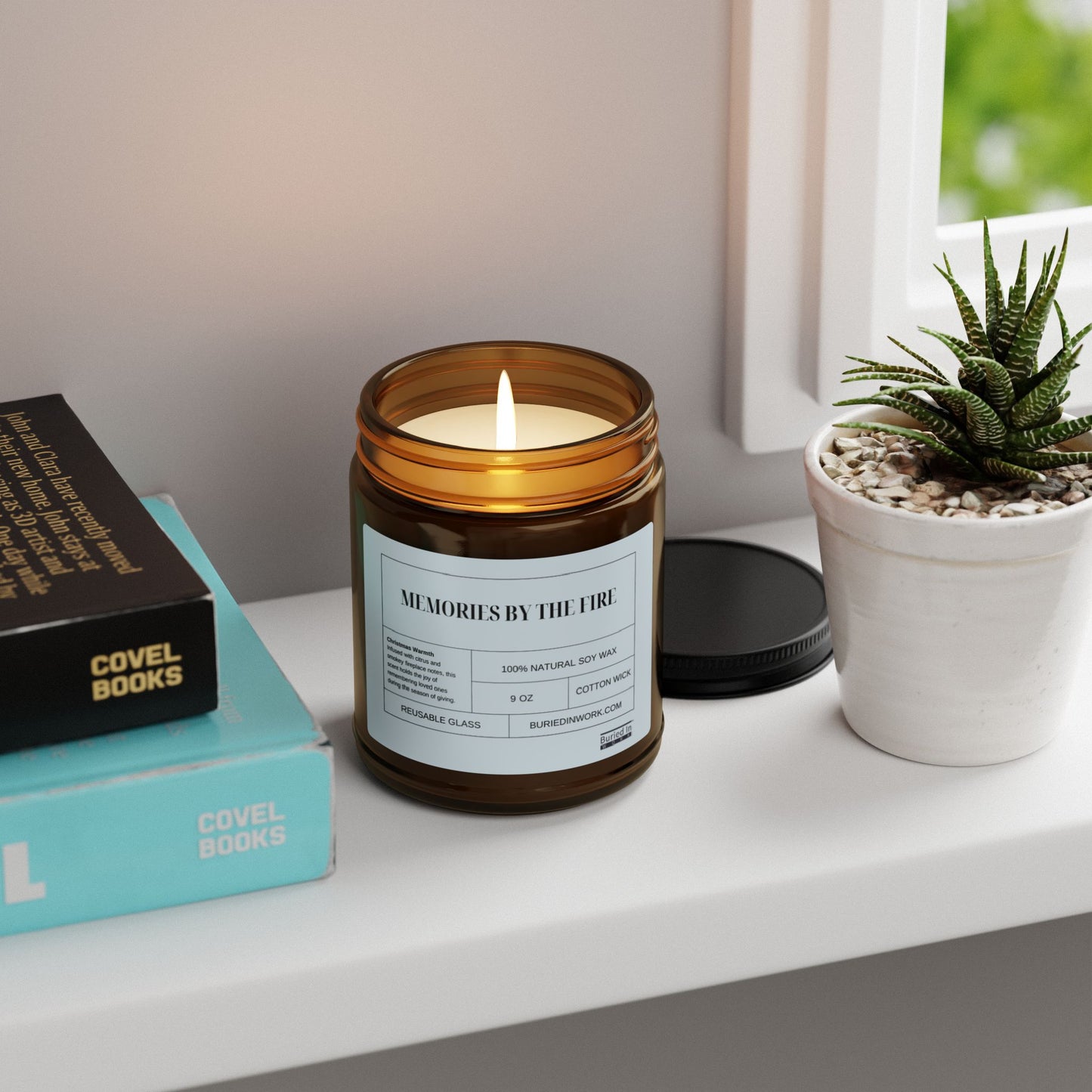 Amber glass soy candle burning on a windowsill beside stacked books and a potted succulent, labelled Memories By The Fire