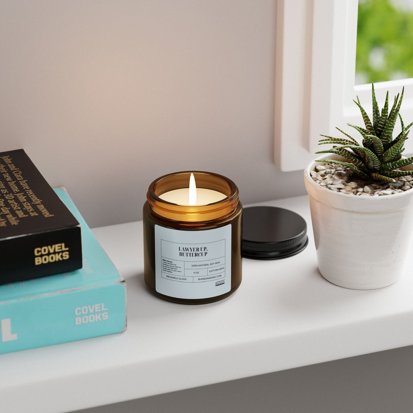 Lit amber glass soy candle on a windowsill beside stacked books and a potted succulent, labelled Lawyer Up Buttercup